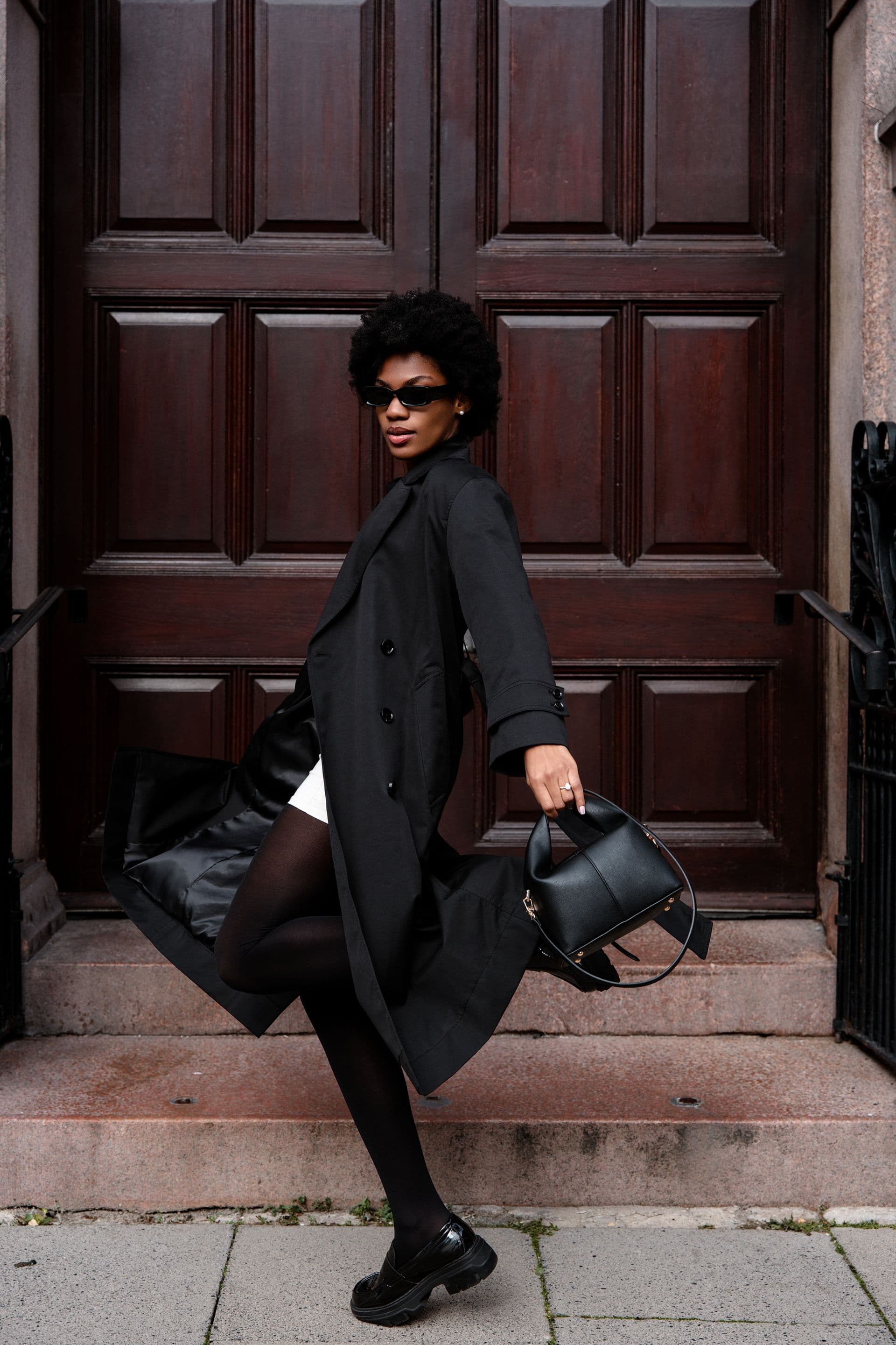 A woman in a long black coat and sunglasses steps down from a dark doorway, a leather bag in hand